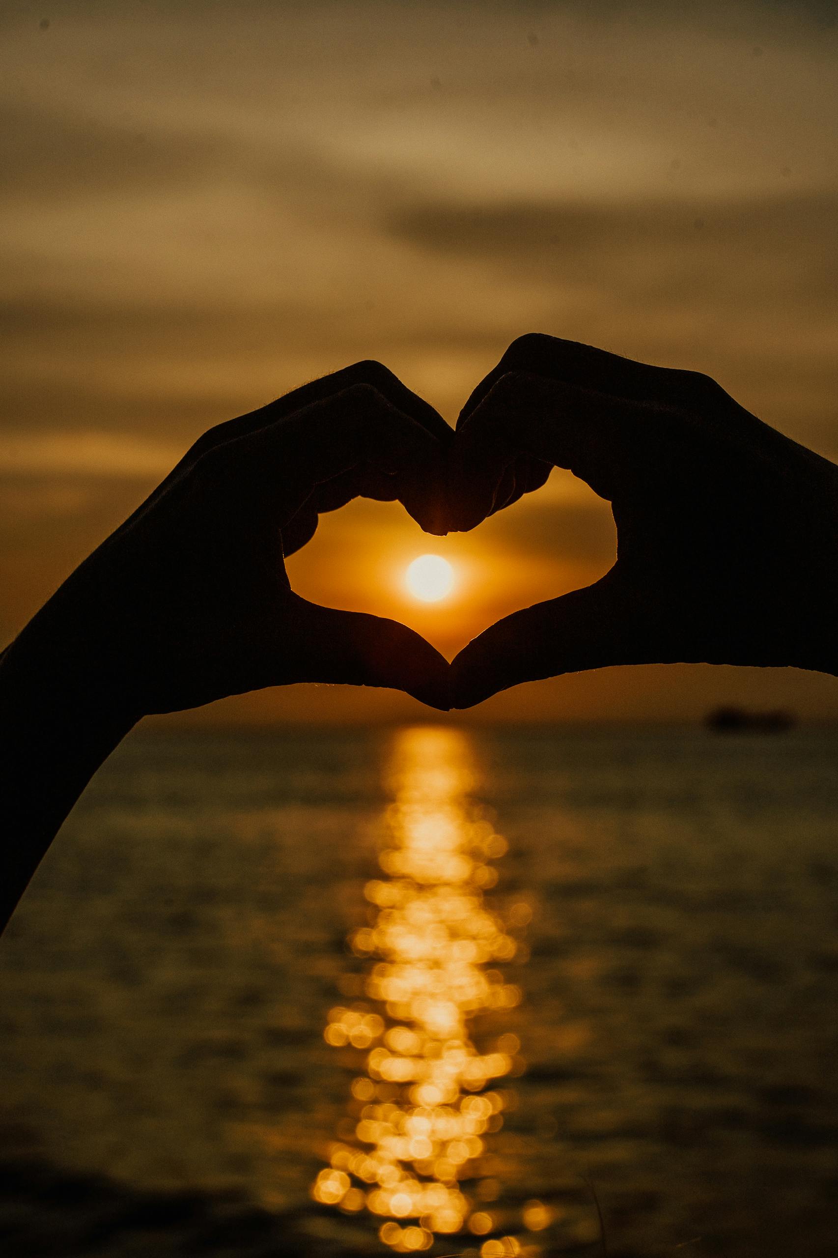 Silhouetted hands form a heart shape against a stunning ocean sunset backdrop.